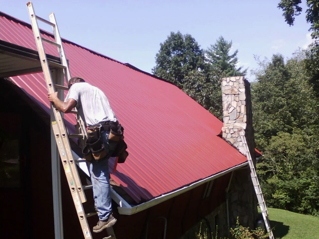 Roofer on a ladder beside a red metal roof and stone chimney — Soffit & Fascia Repair by Roofing Brothers in Cibolo